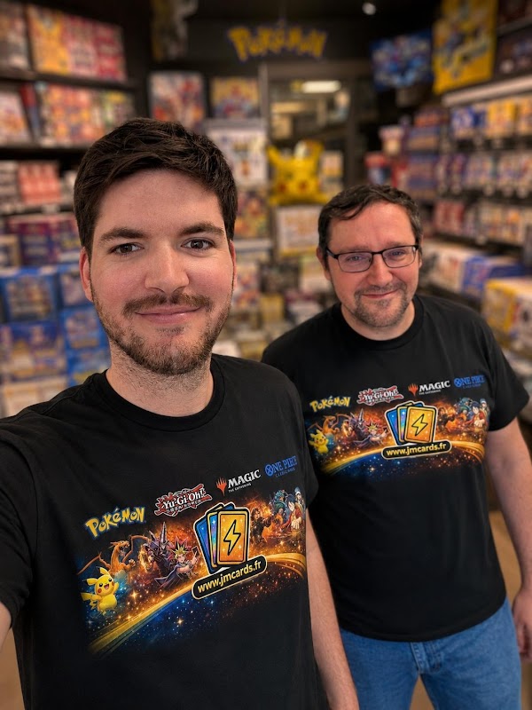 Two men in a game store pose for a selfie, wearing matching black shirts with Pokémon and trading card graphics, shelves of games in the background.